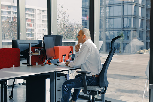 A portrait of a successful and experienced gray-haired mature businessman sitting at a desk in a modern open office. Engrossed in his work, he focuses on the computer monitor, analyzing statistics, commerce data, and marketing plans with precision and expertise. successful, gray-haired, mature, businessman, desk, modern open office, computer, monitor, analysis, statistics, commerce data, marketing plans, expertise