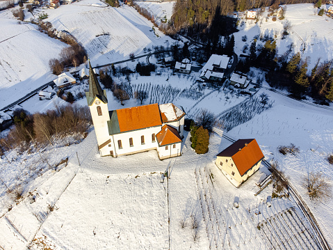 Aerial view of a sun kissed solitary church on a snowy hill with forested mountains in the background during winter.