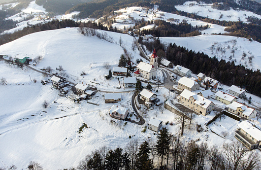 Aerial view of a quaint snow-covered village with a church spire, surrounded by winter trees.