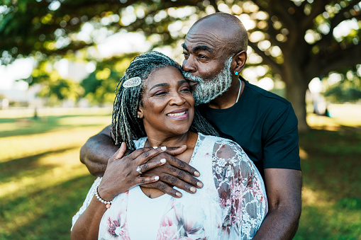 Handsome and active African American senior man affectionately embraces his wife and gives her a kiss on the forehead while enjoying a relaxing afternoon walk through a park in Honolulu, Hawaii during vacation.