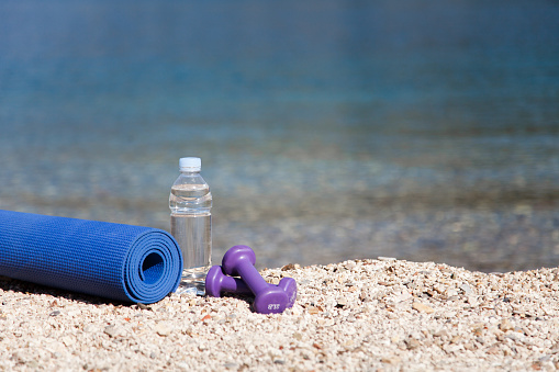 Fitness equipment on sea beach. Sport class in resort. Bottle of clean drinking water, two purple female dumbbells and yoga mat at blue ocean background. Concept of healthy lifestyle and traveling.