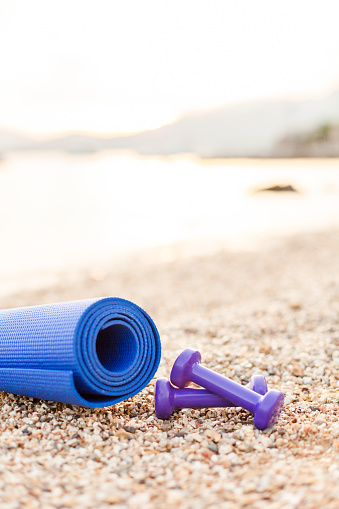 Two purple dumbbells and blue fitness yoga mat on stone and pebble sea beach. Sport background. Concept of healthy lifestyle and traveling. Sport female equipment. Copy space.