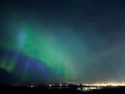 Bright green and purple northern lights (aurora borealis) in the night sky of Reykjavik, Iceland. The sky is filled with light and the city lights are visible on the horizon. There is a reflection on the water. There are small white stars in the sky above. Sightseeing in Iceland.