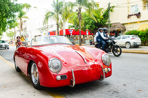 In Playa del Carmen, Mexico a red vintage Porsche is parked along the street with people passing on a scooter and bicycle.