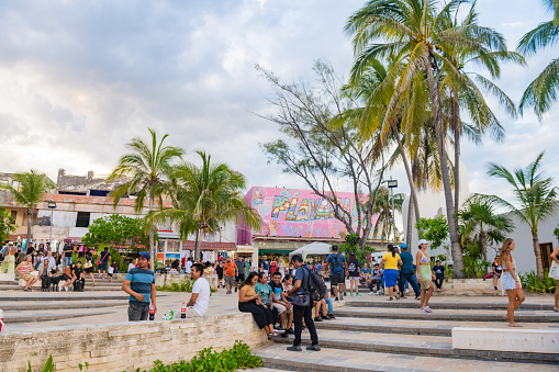 In Playa del Carmen, Mexico people sit outdoors along ledges in a plaza lined with retail shops and palm trees.