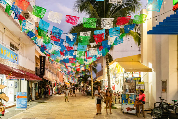 In Playa del Carmen, Mexico traditional flags with cutouts hang above the street lined with shops and people walking at dusk.