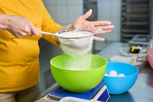 Cropped photo of cook in uniform sifting flour through sieve over bowl placed on kitchen scale on tabletop