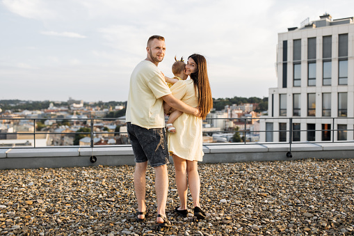 Back view of friendly family of three standing on rooftop with view of city. Beautiful wife standing next to husband and looking at daughter with love. Concept of lifestyles and relationship.