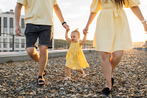 Close up of young couple in love holding hands with cute little daughter making her first walk on summer building terrace. Charming girl in yellow dress feeling support and care from parents.