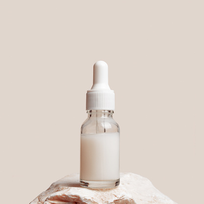 Glass dropper bottle with white liquid essence, serum, cream on stand from natural stone, minimal trend beige background. Natural Cosmetic, monochrome aesthetic mock up cosmetic product for skin care