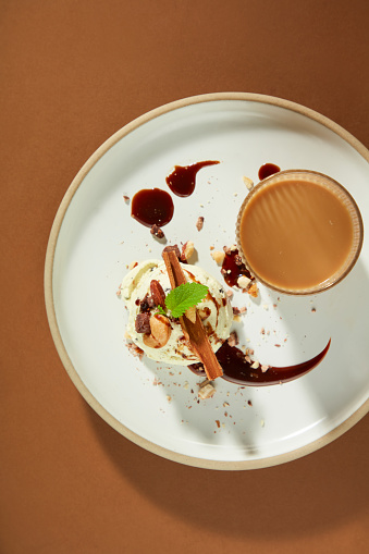 View from above of an elaborately decorated ice cream ball on a plate with crushed almonds, cinnamon, chocolate sauce and mint leaves, next to a glass of milky coffee. Brown background.