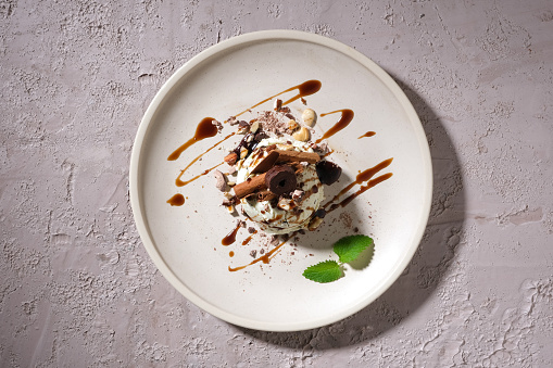 Top view of an ice cream decorated with chocolate sauce and crushed nuts on a white ceramic plate on a rough concrete surface. Food advertising.