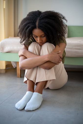 Young African American girl sits with hands on knees. Stress emotion, vertical image.