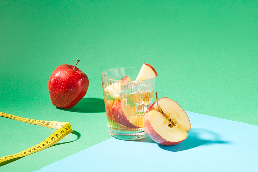 On a green backdrop, detox water, an apple, tape measure, and a blue paper sheet. Nutrient-packed apples, low in calories, support weight loss. Embrace a refreshing journey to wellness.
