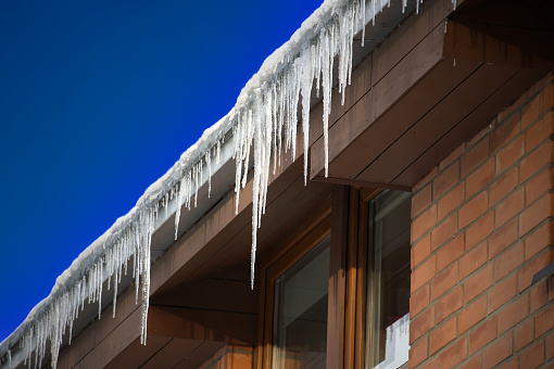 Lineup of large icicles hanging from the edge of a brown wooden residential house's roof