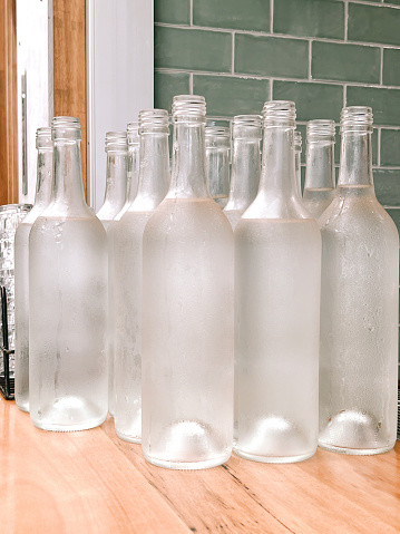 Vertical closeup photo of a group of clean, clear glass wine bottles, used as water bottles on cafe tables, on a wooden bench next to a tiled and timber wall, in a cafe restaurant.