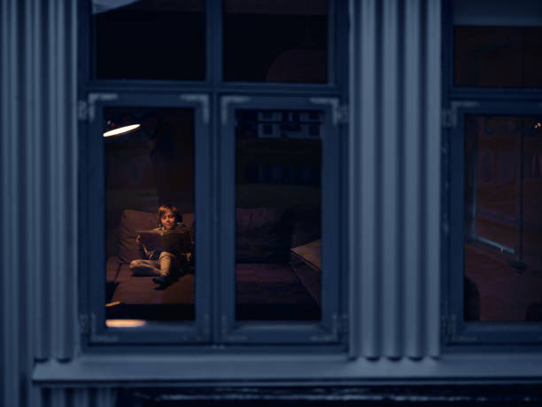 Smiling little boy reading a book while relaxing on sofa in the living room at night. The view is through window. Photographed in medium format.