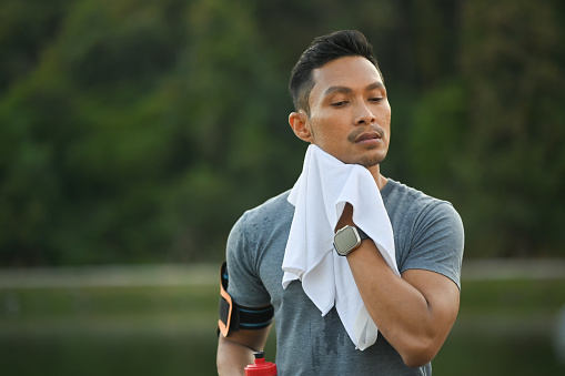 Young muscular man resting and wiping sweat with a towel after morning cardio workout in park.