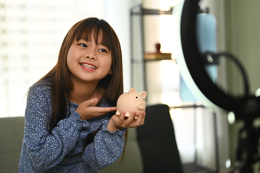 Pretty asian girl holding piggybank in front of camera while recording video on smartphone. Saving money for future concept.