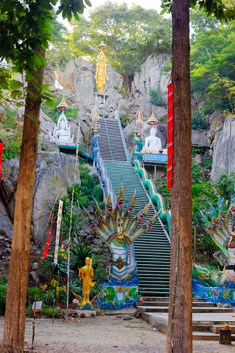 Steps up to buddha flanked by thai Nakhon snakes and merman and mermaid on grounds of temple in small village Paranghmee near Noen Maprang in Phitsanulok province. View between trees and meditation area. Slim golden Buddha is at end of steps