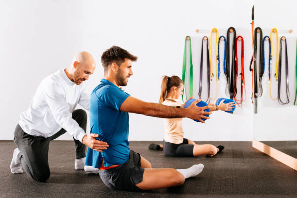 istock Instructor assisting young male and female doing stretch exercise with foam roller at gym 1916711977