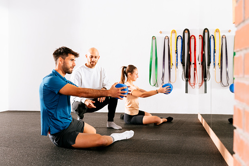 Side view of young man and woman in activewear sitting on floor while doing exercises with elastic rubber equipment during fitness training at modern studio