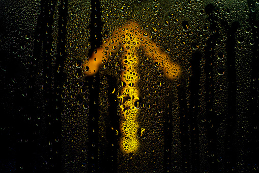 Glowing arrow sign pointing up behind wet glass. Raindrops on the window. Background on the theme of positive change and improvement.