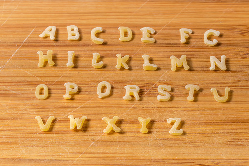 Raw pasta in the shape of capital letters of English alphabet placed in rows in the alphabetical order on the bamboo cutting board, close-up