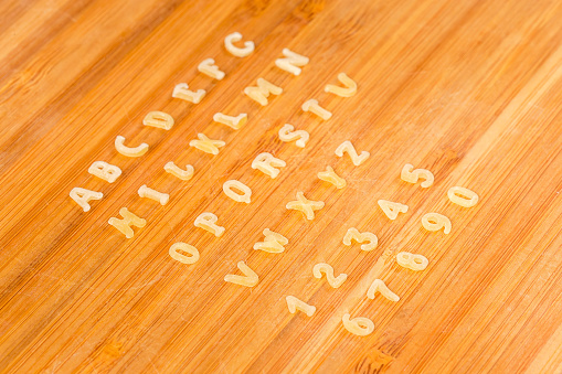 Raw pasta in the shape of capital letters of English alphabet and Arabic numerals placed in rows in the alphabetical order on the cutting board, side view close-up in selective focus