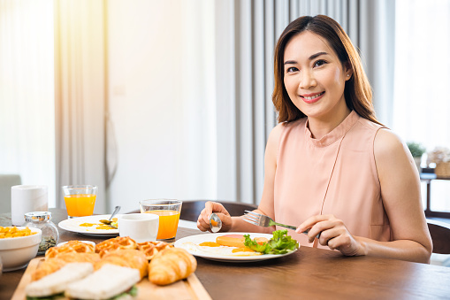 Asian young woman sitting kitchen table food having eating healthy breakfast at home, female eat fresh breakfast served food with beverage before go to work in the morning at home