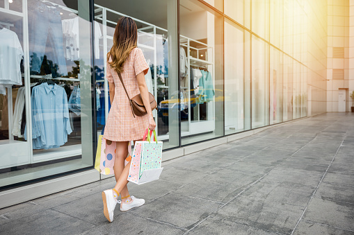 A beautiful woman carrying shopping bags walks past a clothing store window display, holding a gift bag and enjoying the fun of shopping in the mall. Retail therapy is a glamorous lifestyle.