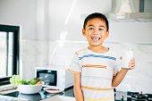 Portrait of cute Asian little boy in kitchen holding milk. Smiling son enjoys drink, radiating joy. Happy preschool child sips calcium-rich liquid, feeling cheerful at home give me.