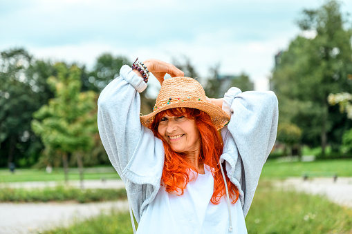 Cheerful senior woman with long red hair and casual clothes posing with her straw hat on the head in a public park in summertime.