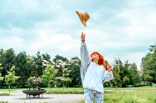 Cheerful senior woman with long red hair and casual clothes throwing her straw hat in the air in a public park in summertime.