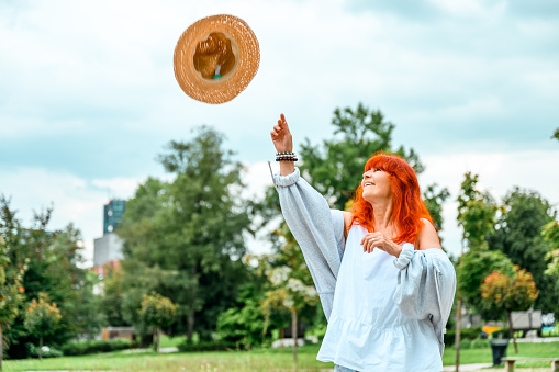 Cheerful senior woman with long red hair and casual clothes throwing her straw hat in the air in a public park in summertime.