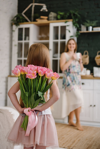 Happy birthday. Mom with daughter surprised. Kid congratulates mother and gives bouquet of tulip flowers in kitchen at home. Mother's Day concept. Greeting card. Women's Day. Back view. Family holiday