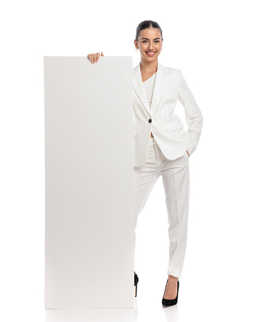 confident woman in white suit showing and presenting empty board while standing with hand in pocket and smiling on white background