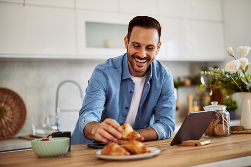 A young adult male freelancer smiles and takes a croissant for a snack on a break while leaning on a kitchen counter in front of a tablet for his work.