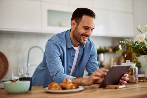 istock A cheerful man answers a video call on a tablet while leaning on a kitchen counter. 1916794280