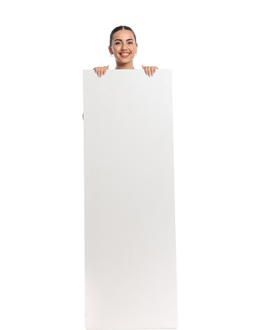 smiling woman holding empty board, covering body and being happy while looking forward on white background