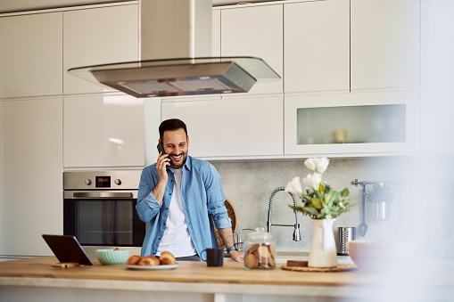 A cheerful adult househusband laughing and talking with his wife over a mobile phone in a kitchen setting.