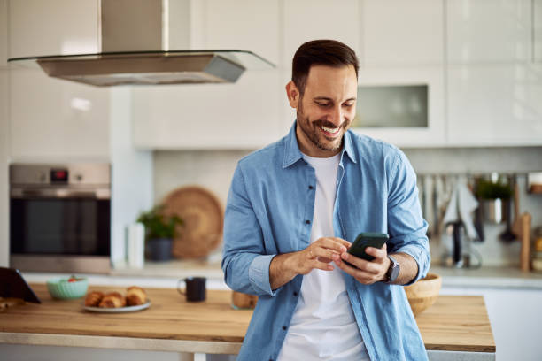 A smiling freelance man sending his coworker a funny text message while standing in a kitchen.
