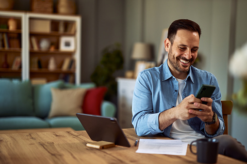 A cheerful man texting his friends on his cellphone while taking a break from his remote work on tablet and sitting in a coffee shop.