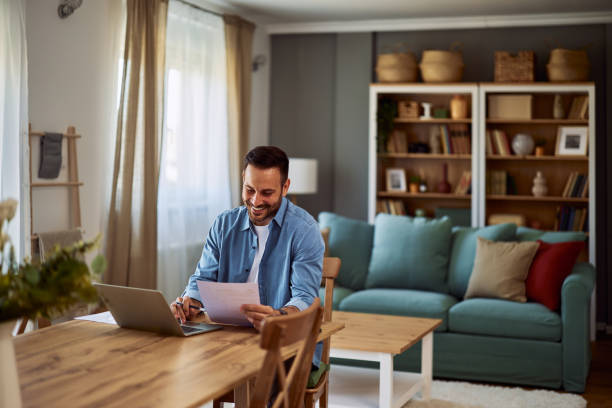 istock A smiling male remote data analyst using a laptop and looking at data on a paper in front of him while sitting a table. 1916802434