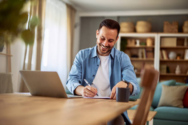 un giovane insegnante maschio adulto sorridente che valuta i test dei suoi studenti mentre è seduto a un tavolo davanti a un laptop. - grading teacher desk writing foto e immagini stock