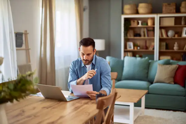 Photo of A focused male remote worker analyzing data on a paper he is holding while sitting at a desk in front of a laptop.