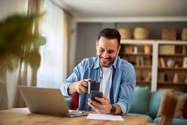 A happy young adult remote worker holding a cup of coffee while scrolling through his phone and sitting in front of a laptop on his work break.