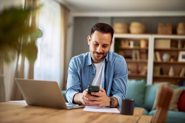 istock A smiling man checking his messages on his smartphone while sitting at a table with a laptop in front of him. 1916805103