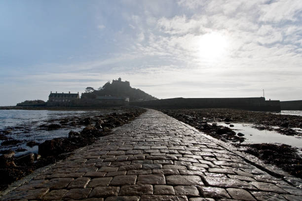 Marazion, Cornwall, England, UK: St Michael's Mount - tidal island on the southwestern tip of England, lying south the town of Marazion in Cornwall. It can be reached either by ferry or, at low tide, via a narrow causeway from Marazion. Historically, St Michael's Mount was a Cornish counterpart of Mont-Saint-Michel in Normandy, France, which is also a tidal island, and has a similar conical shape, though Mont-Saint-Michel is much taller - Cornwall Area of Outstanding Natural Beauty