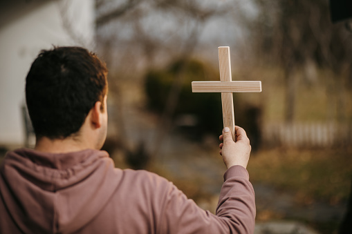 Close-up shot of man holding religious wooden cross and praying in front yard